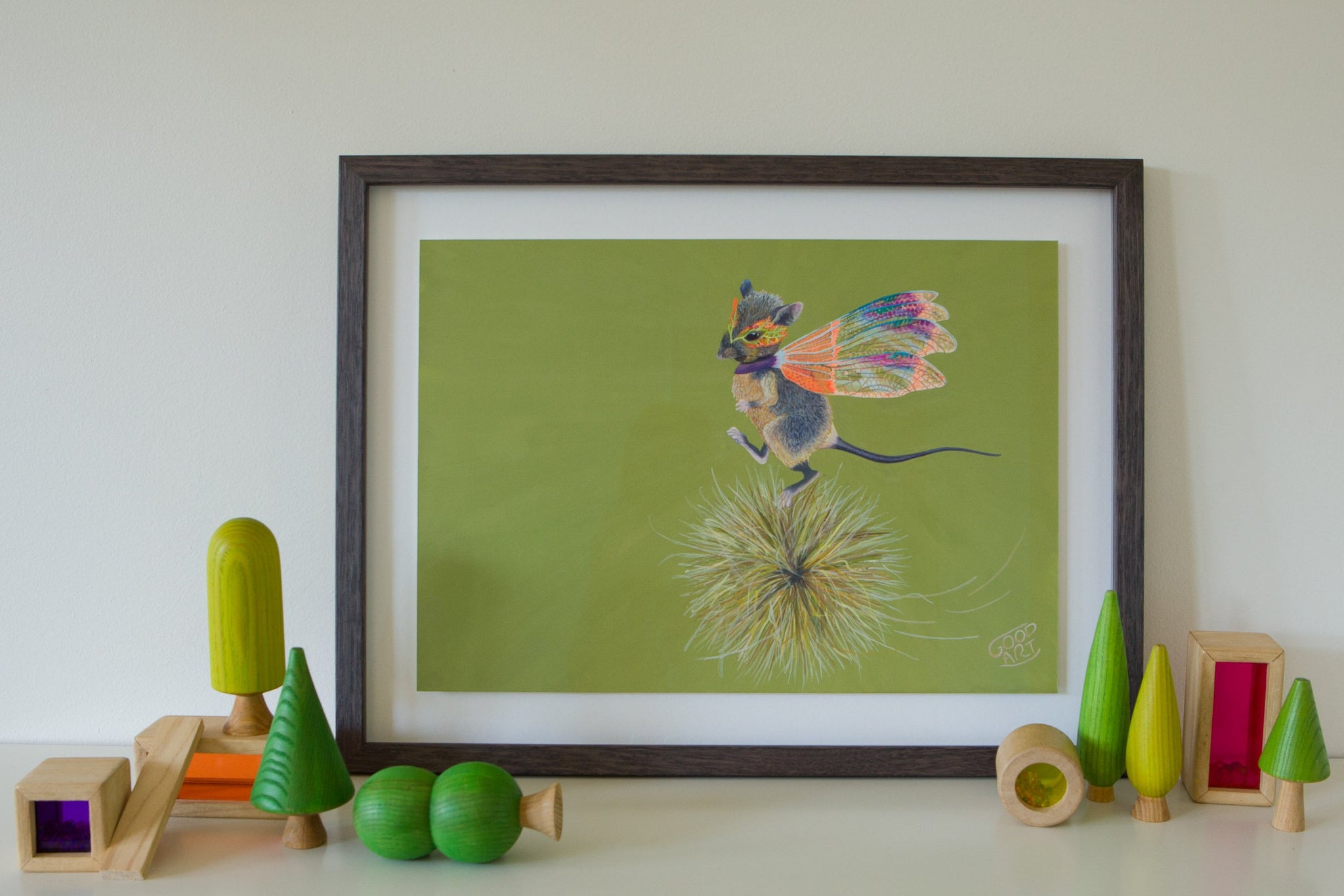 Western Australian marsupial, the Dunnart. Mouse-like in appearance. In this artwork the Dunnart wearing a set of dragonfly wings runs along on top of a spinning spinifix. A predominately olive coloured background. Artwork sits on shelf amongst wooden kids toys. Kids prints painted by artist Jaelle Pedroli.