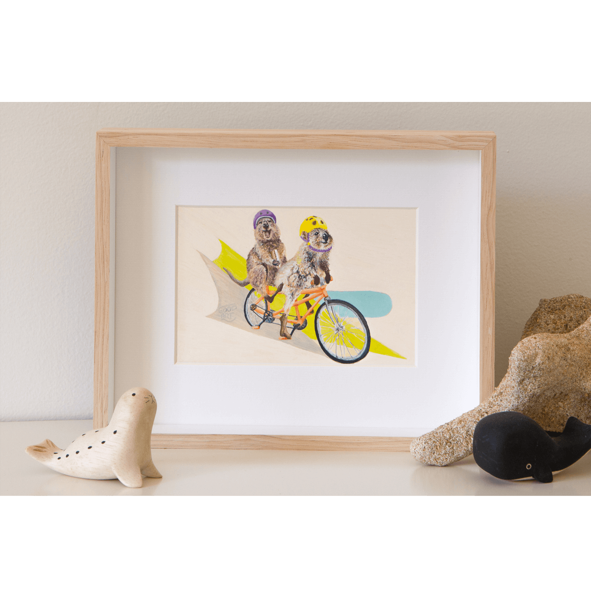 an artwork of two Rottnest quokkas riding a tandem bike. The laughing quokka at the back with legs in the air isn’t peddling but instead is eating a cream bun. Framed original painting of quokkas on a shelf.