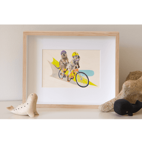 an artwork of two Rottnest quokkas riding a tandem bike. The laughing quokka at the back with legs in the air isn’t peddling but instead is eating a cream bun. Framed original painting of quokkas on a shelf.