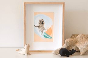 Quokka surfing a barrel wave with the sunsetting behind her. Green surfboard, orange sky. Framed original artwork sitting on a shelf. 