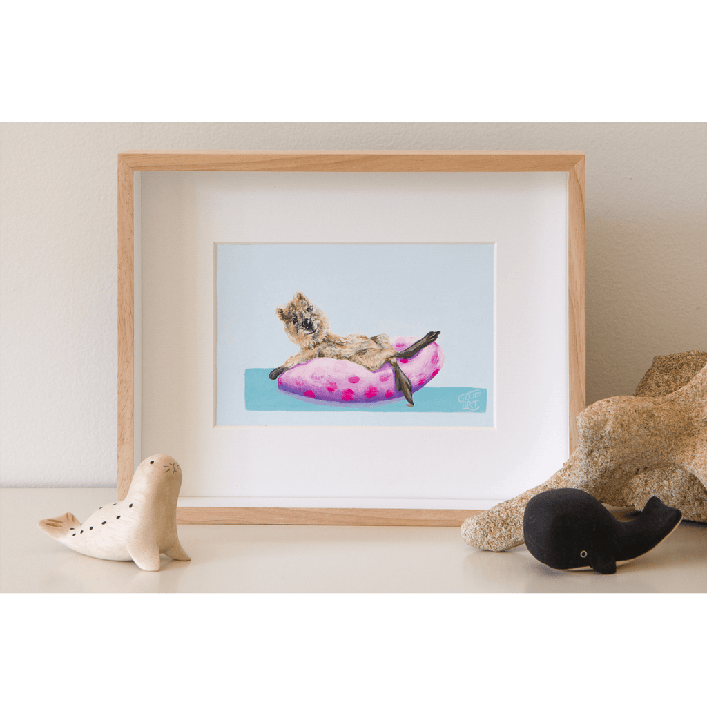 An original painting of a Rottnest Island quokka reclining on a pool doughnut, whilst dipping her toe into the sea. Pink and purple spotted doughnut. The artwork is framed and sits on a shelf. Artwork painted by Australian artist Jaelle Pedroli for Good Art.