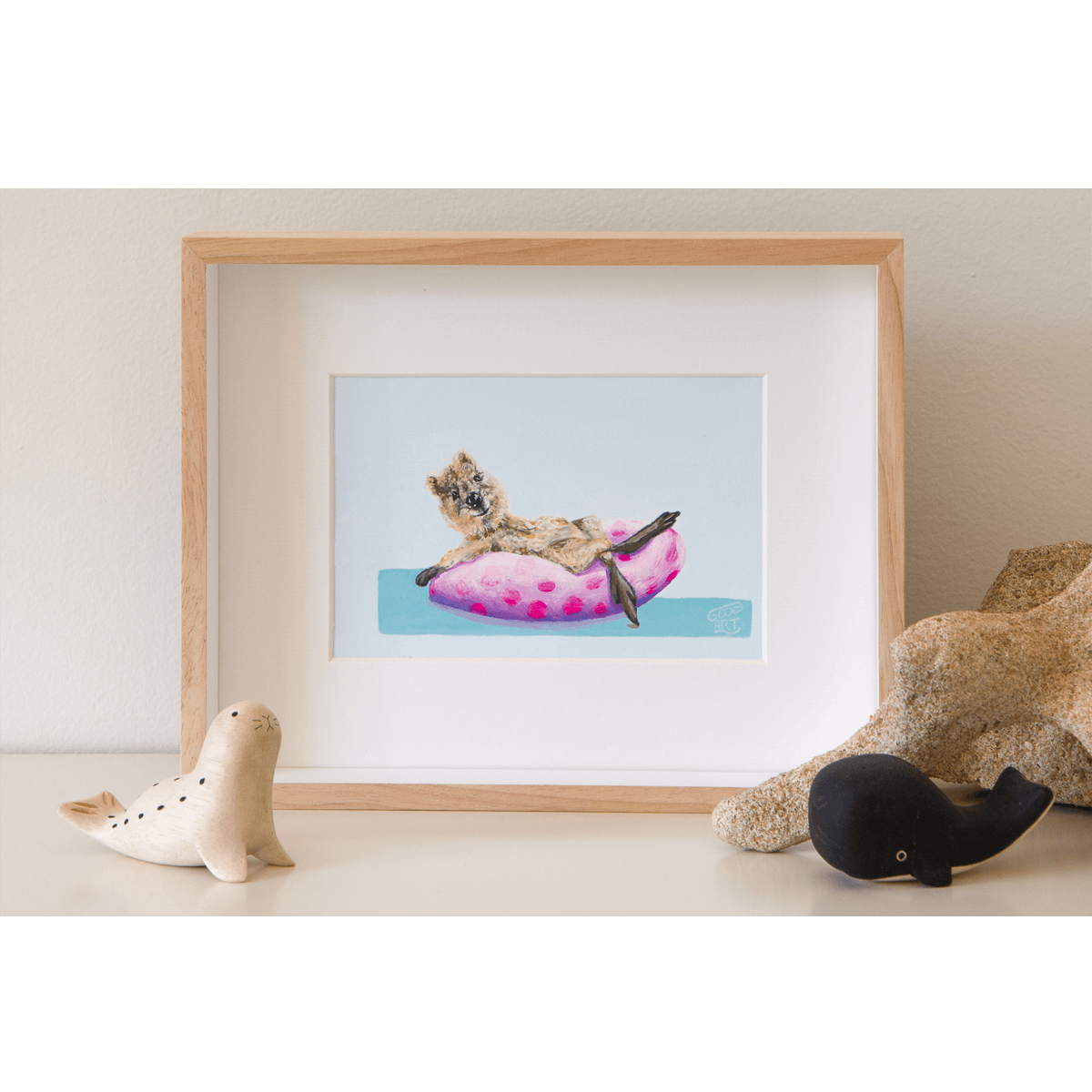 An original painting of a Rottnest Island quokka reclining on a pool doughnut, whilst dipping her toe into the sea. Pink and purple spotted doughnut. The artwork is framed and sits on a shelf. Artwork painted by Australian artist Jaelle Pedroli for Good Art.