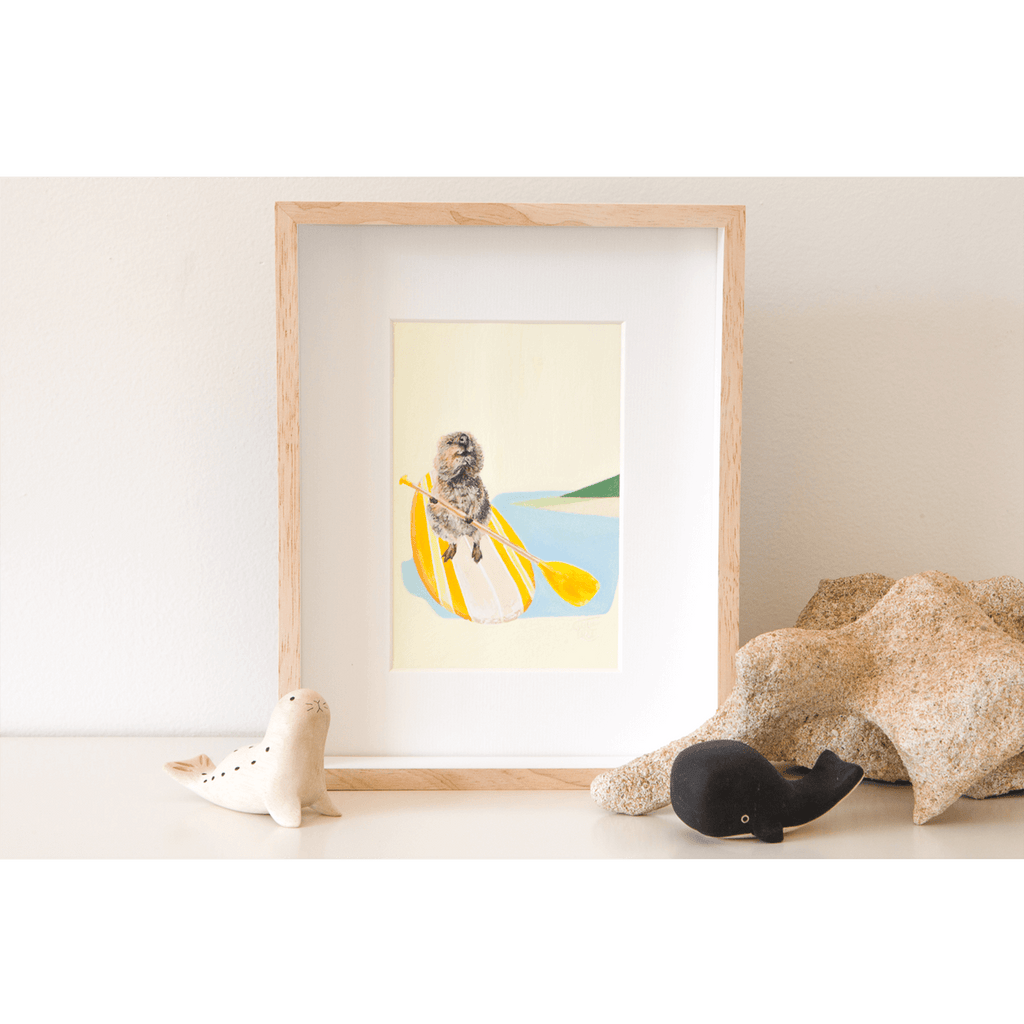 An original painting of a Rottnest quokka blissfully paddling a yellow stand up paddle board over the ocean. Frames original artwork sitting on a shelf.