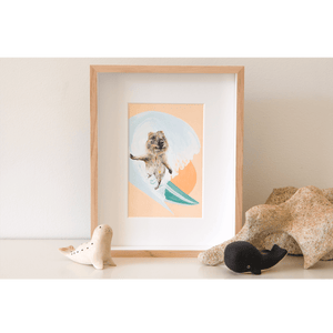 Quokka surfing a barrel wave with the sunsetting behind her. Green surfboard, orange sky. Framed original artwork sitting on a shelf. 